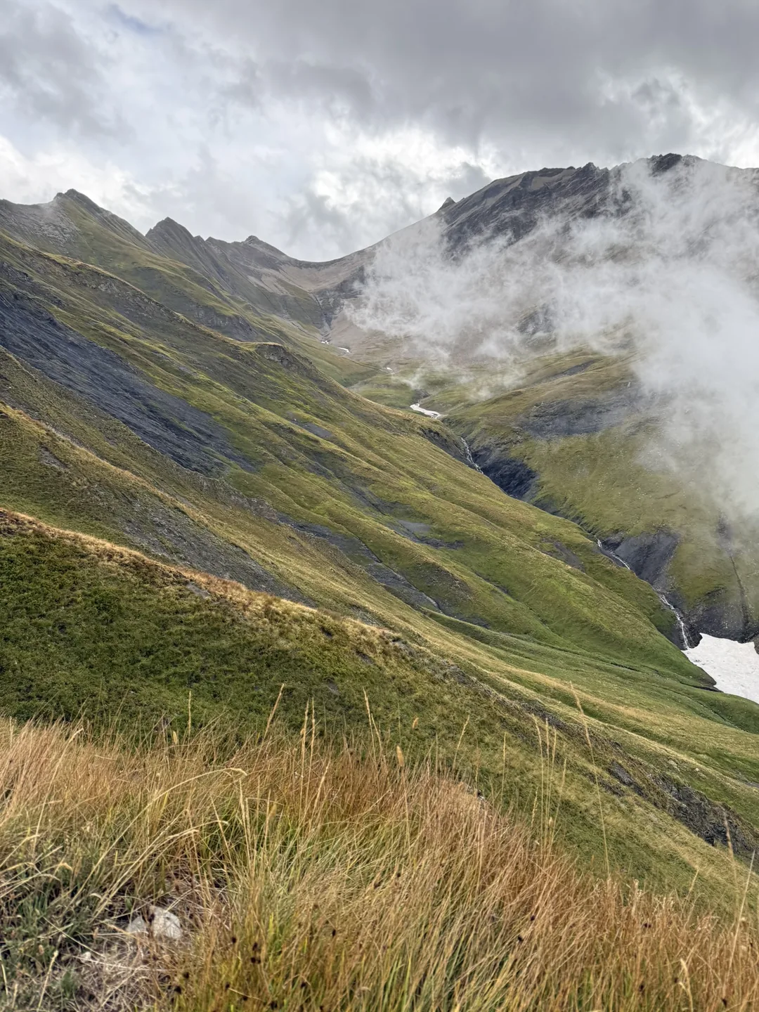 Green alpine flanks with cloud spilling over the ridgeline above a steep gully.