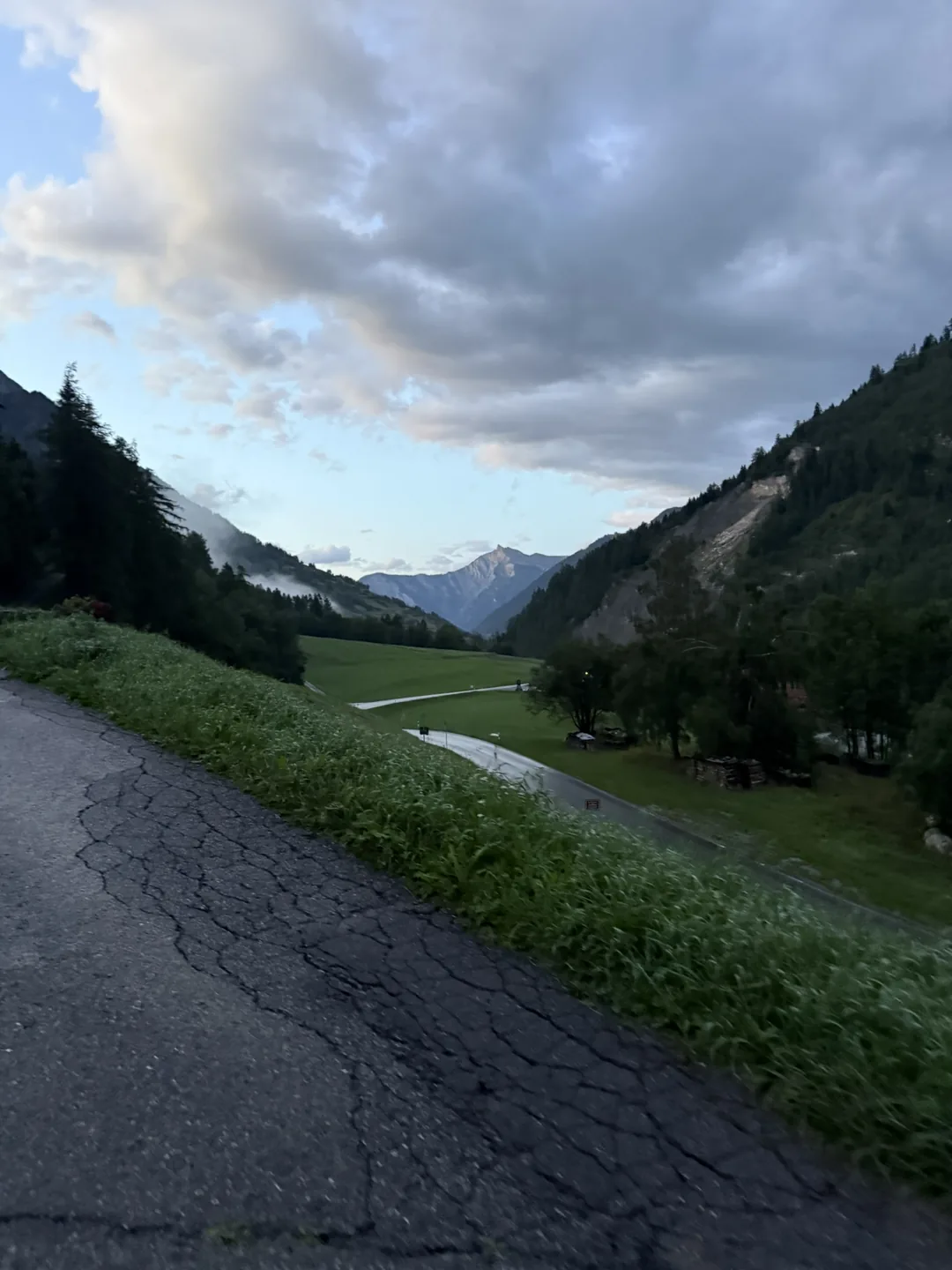 A wet mountain road at dusk running down a green Swiss valley under breaking cloud.